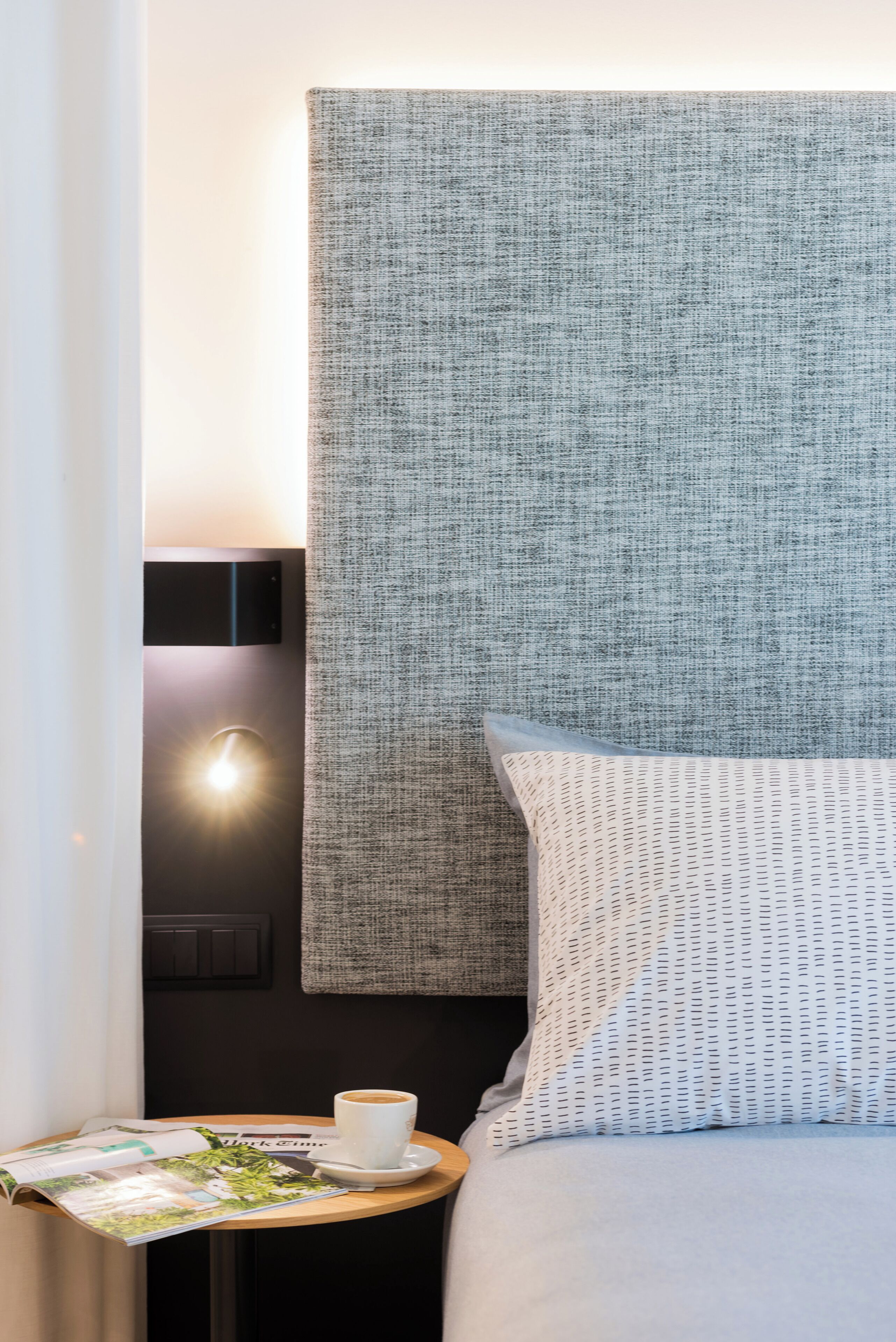 A neatly made bed with a patterned pillow, a small round table with a cup of coffee and a magazine, and a wall-mounted light.