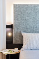 A neatly made bed with a patterned pillow, a small round table with a cup of coffee and a magazine, and a wall-mounted light.