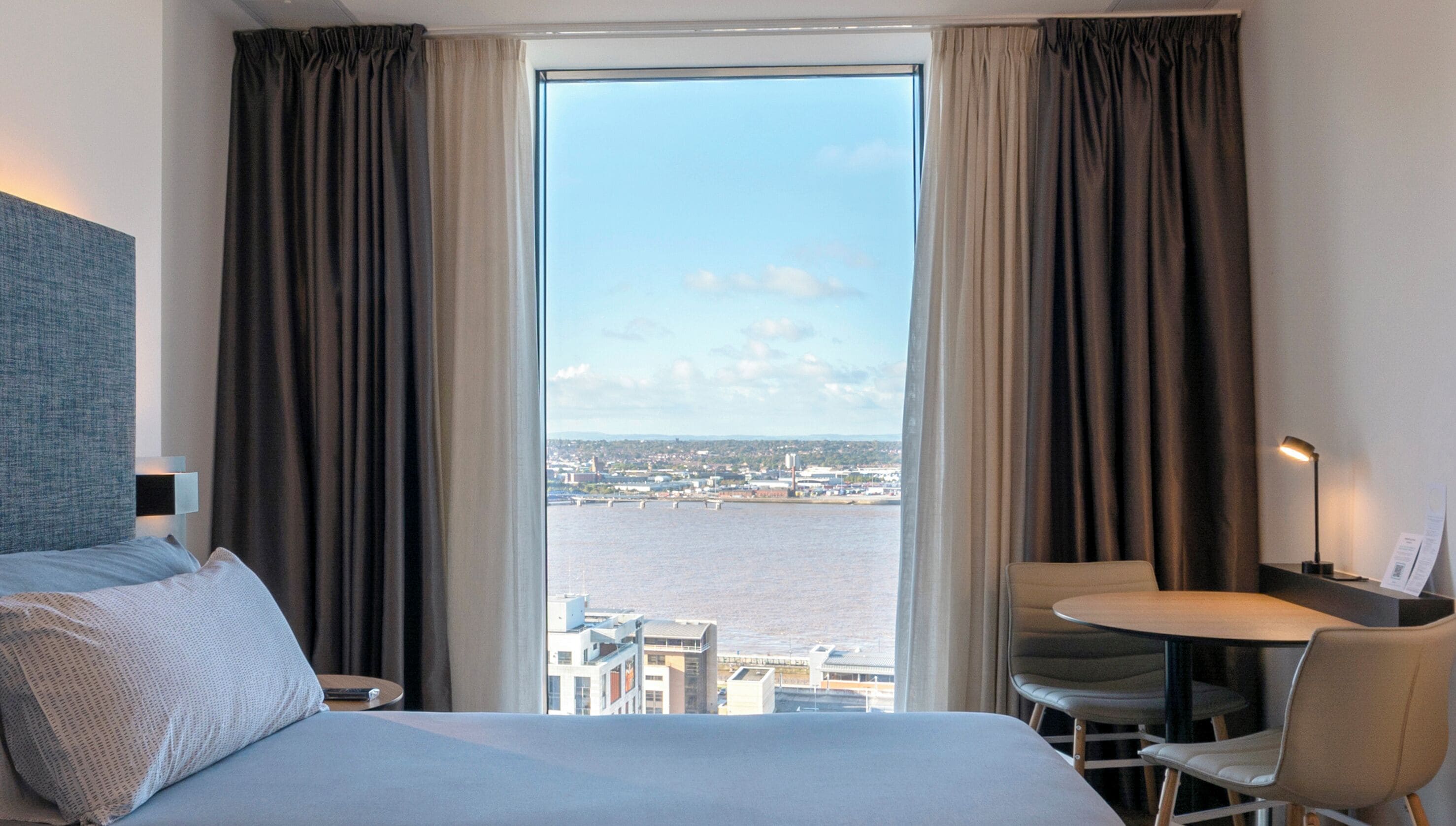 Habitación de hotel con una cama grande, una mesita con una silla, una lámpara de escritorio y vistas a un paisaje urbano a través de un ventanal amplio.