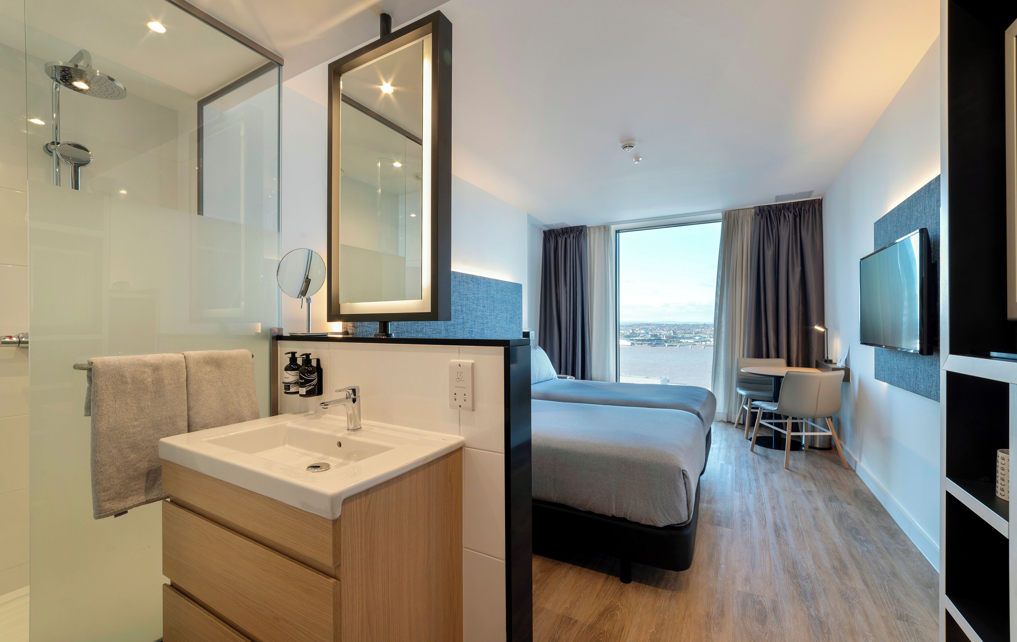 A modern hotel room with a large bed, a desk, a TV, a bathroom with a sink and mirror, and a view of the city.