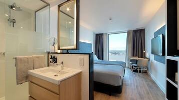 A modern hotel room with a large bed, a desk, a TV, a bathroom with a sink and mirror, and a view of the city.