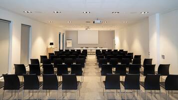 Un salón de conferencias con filas de sillas negras orientadas hacia una pantalla de proyección, un atril y una pizarra.