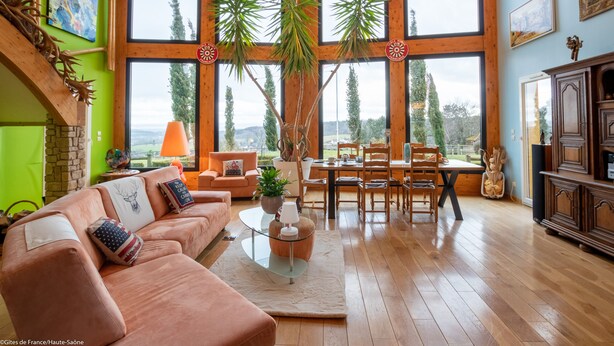 A spacious living room with a large window, a wooden floor, a sofa set, a dining table, and a fireplace. - Au Domaine du Cerf (Vyans-le-Val)