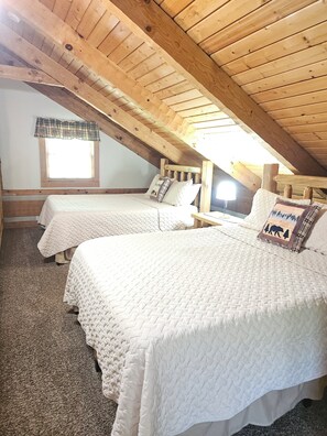 Une chambre confortable située sous les toits, avec deux lits, des poutres apparentes, une fenêtre agrémentée d’une lambrequin à carreaux et une petite lucarne.