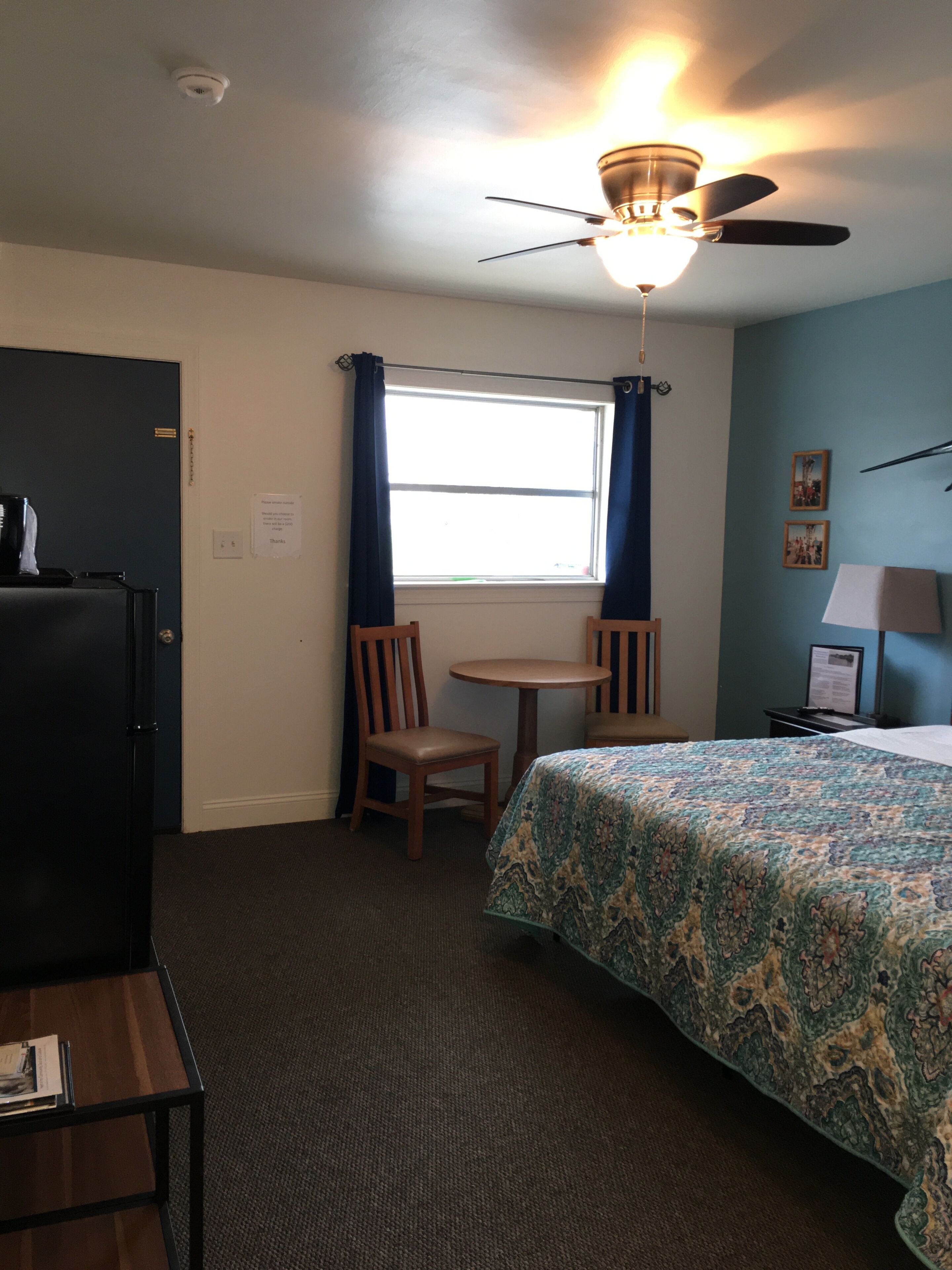 Une chambre d’hôtel comprenant un lit, un bureau, une chaise, une fenêtre avec des rideaux, un ventilateur de plafond et un réfrigérateur.
