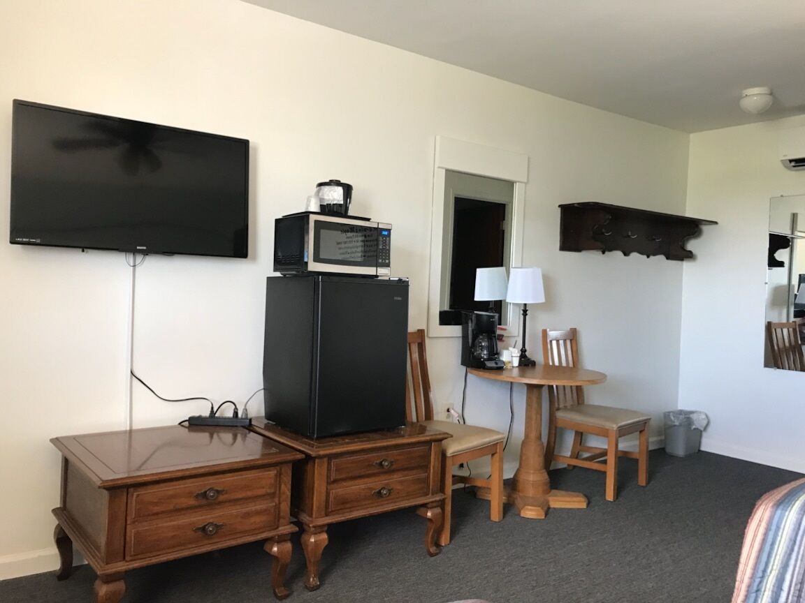 Una habitación con televisión de pantalla plana, microondas, refrigerador, escritorio de madera, silla, una pequeña mesa con lámpara y espejo.