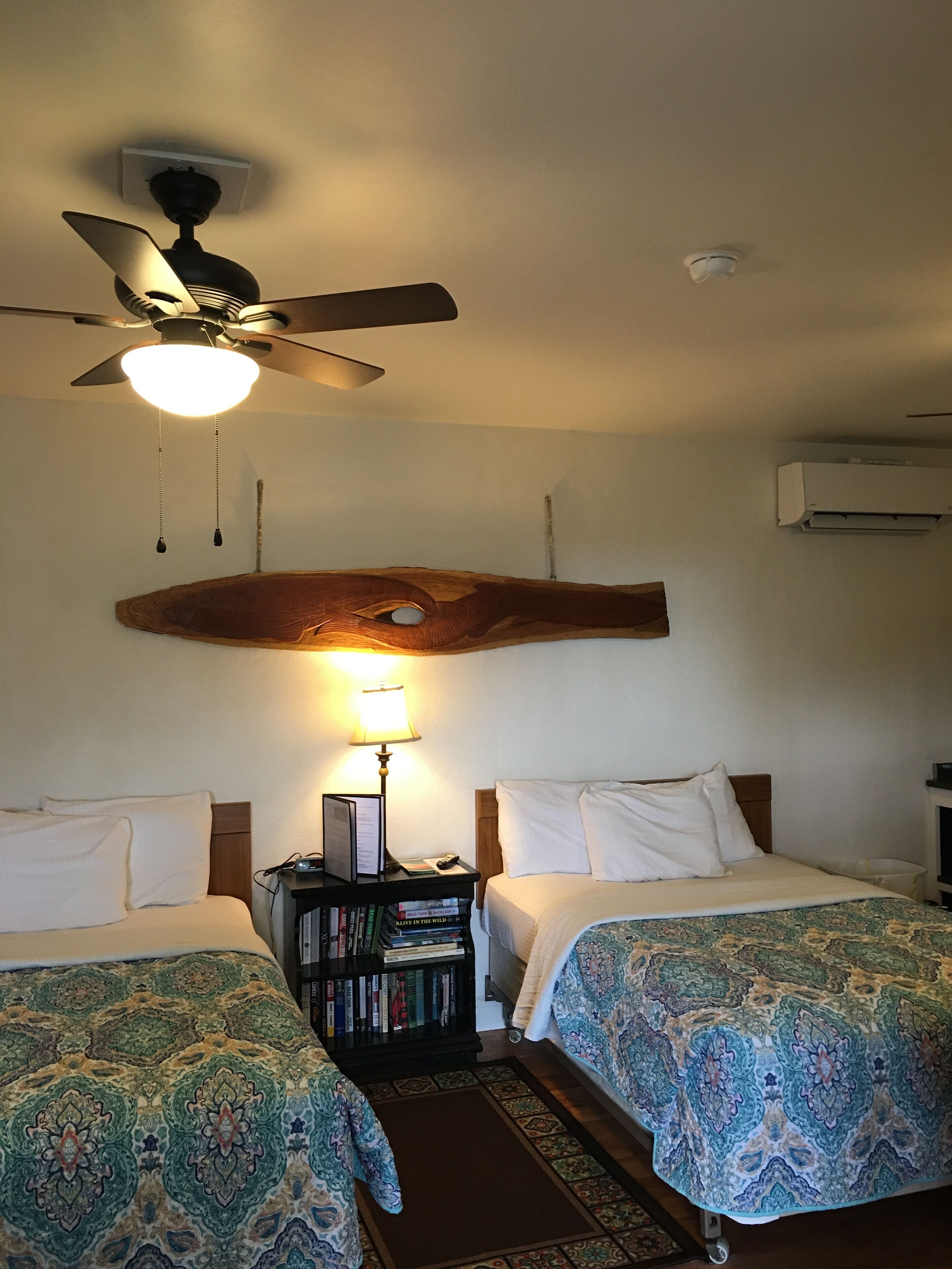 A hotel room with two beds, a ceiling fan, a nightstand with books, and an air conditioning unit.