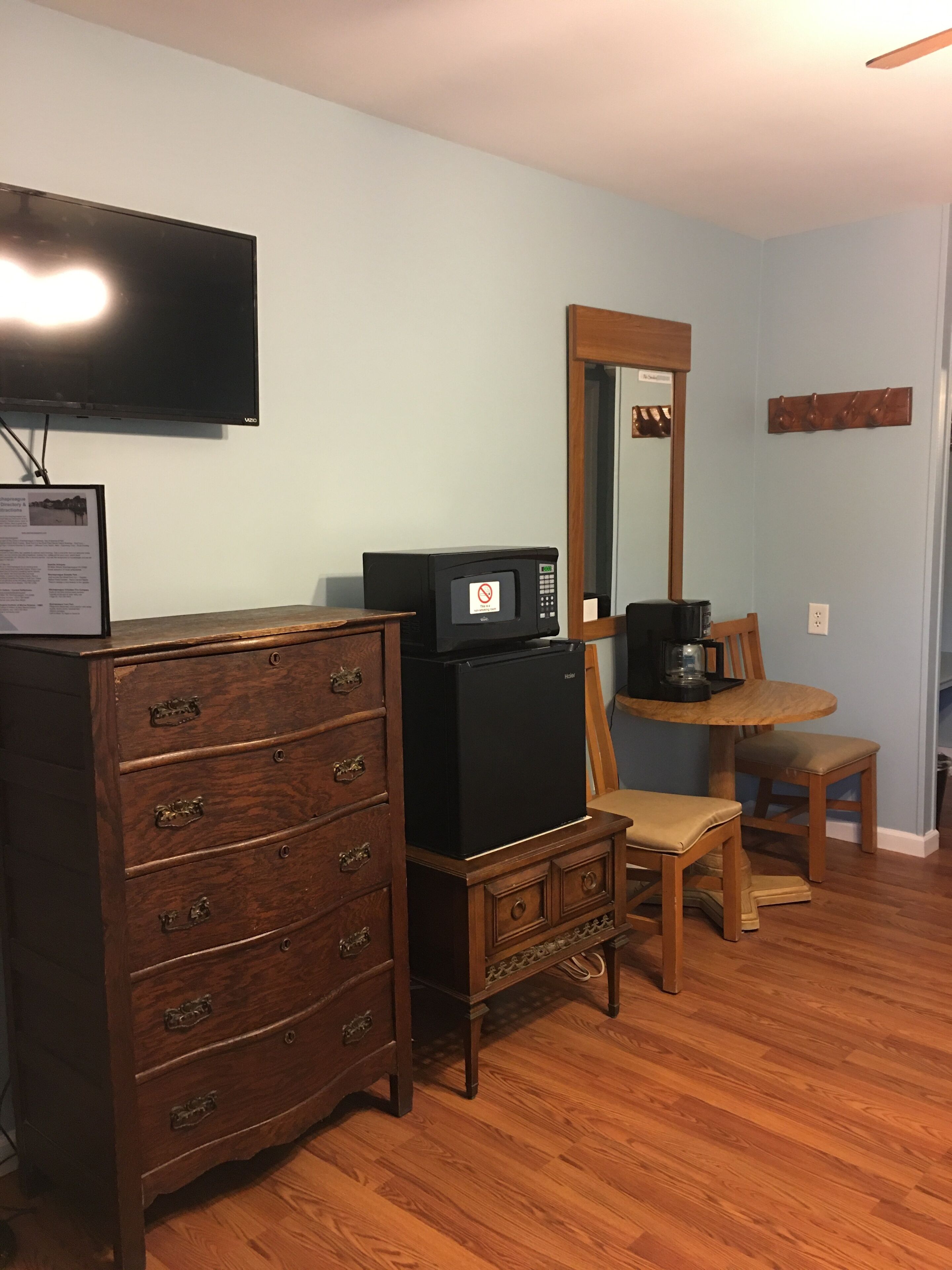 Une pièce comprenant une commode en bois, un réfrigérateur noir, une chaise en bois, une table avec une machine à café et un téléviseur fixé au mur.