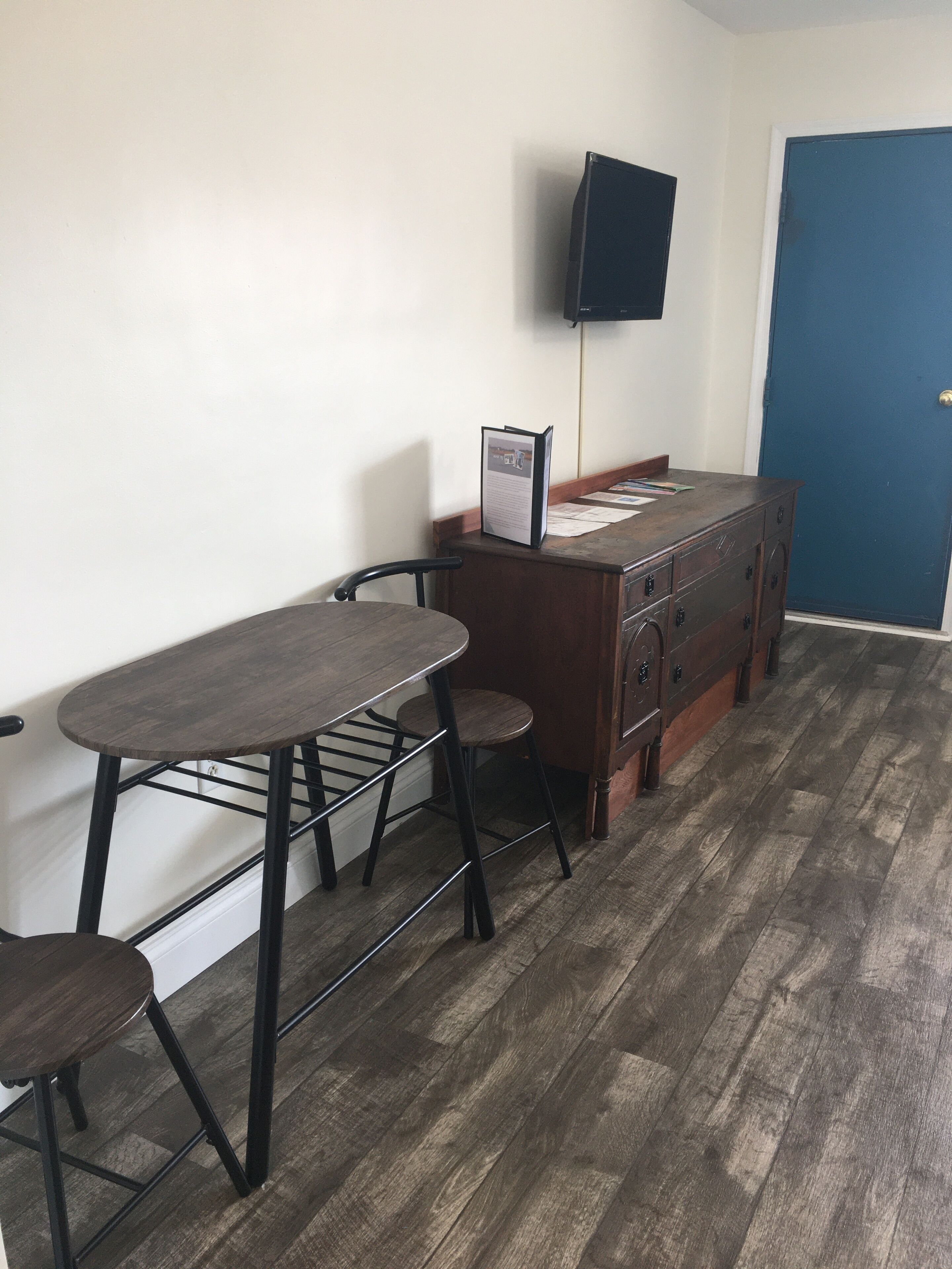 Une pièce avec une table en bois et deux chaises, un bureau avec des tiroirs, un téléviseur fixé au mur et une porte bleue.