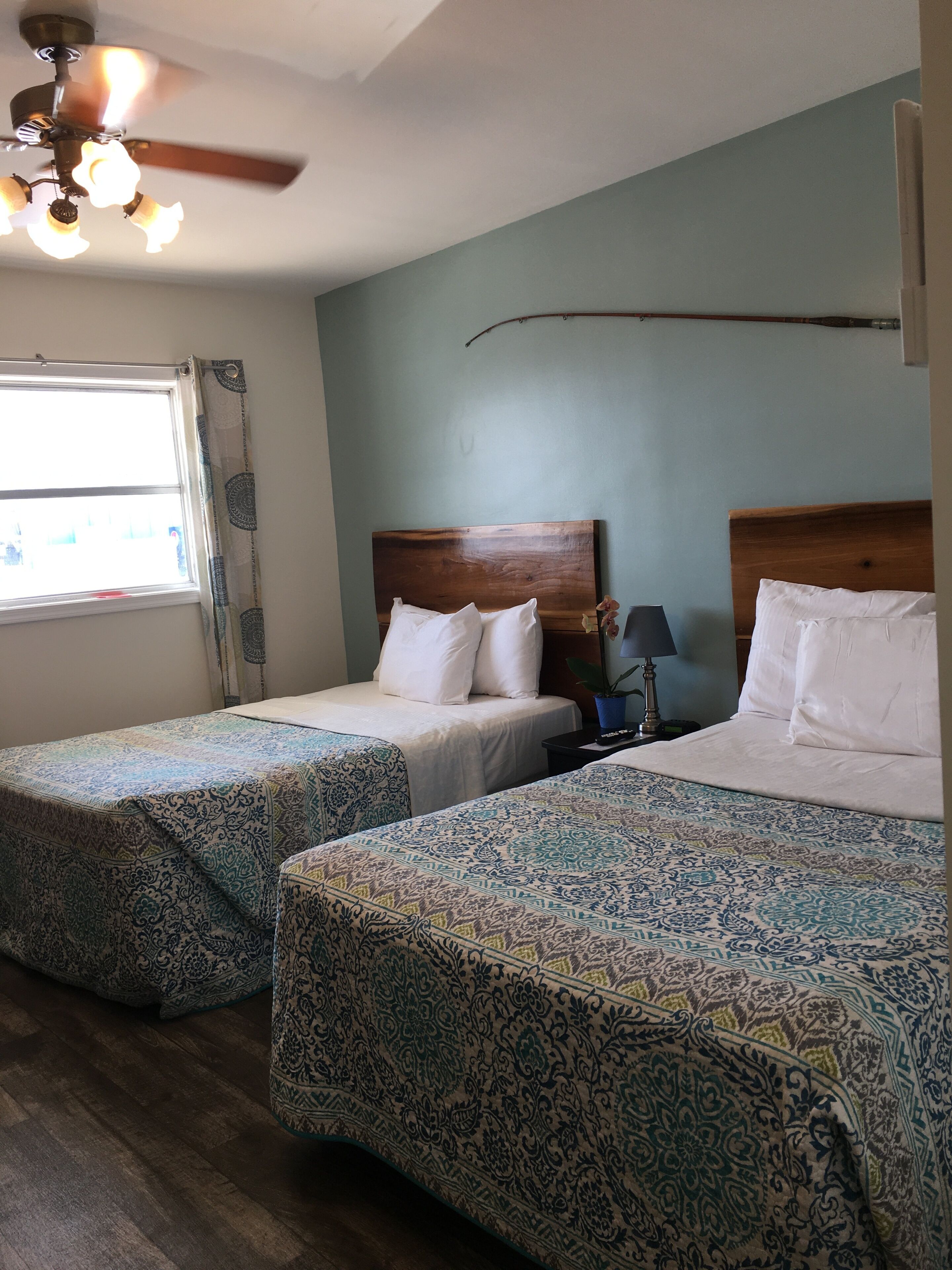 A hotel room with two beds, a ceiling fan, a window with curtains, and a nightstand with a lamp.