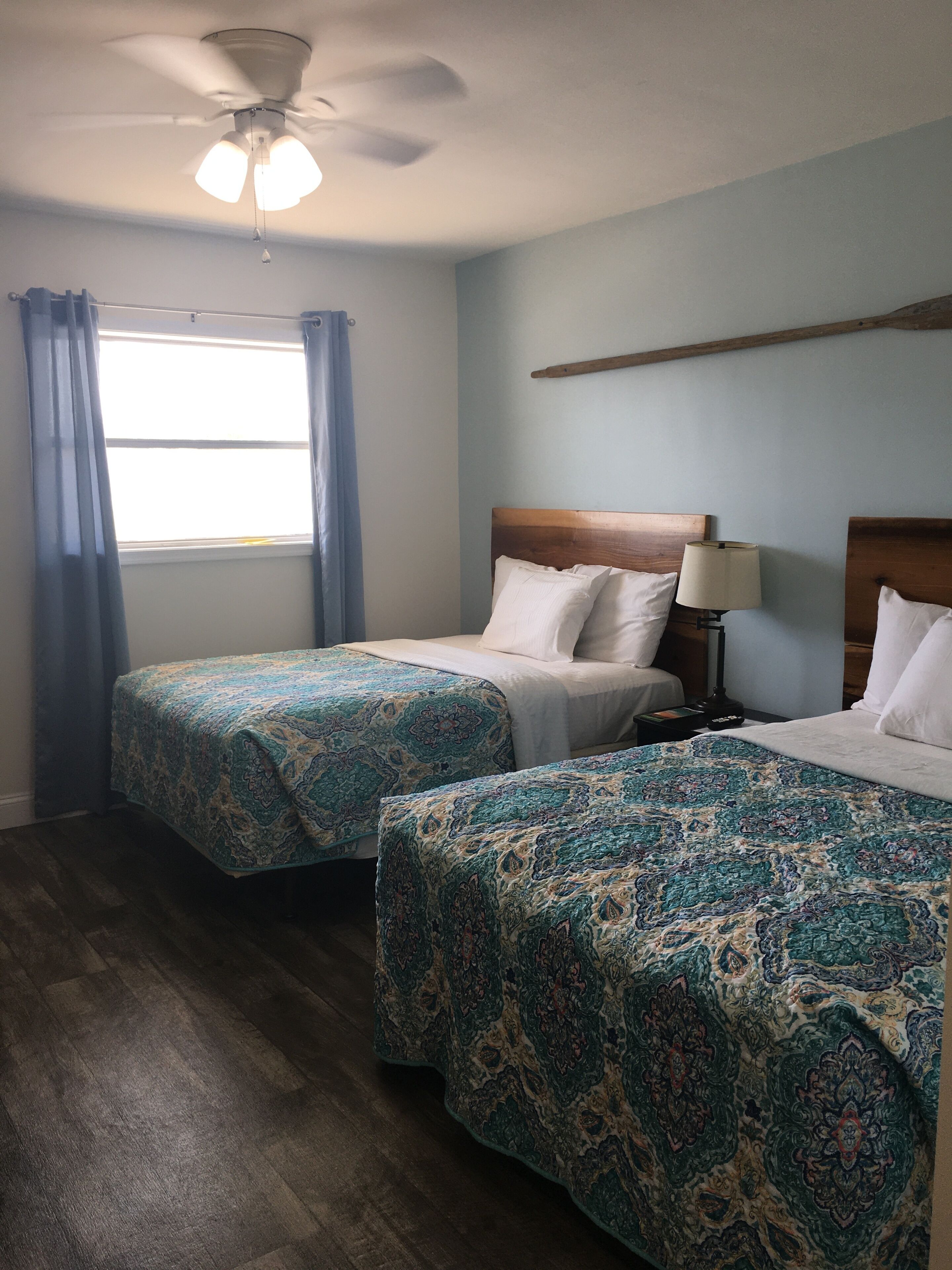 A hotel room with two beds, a ceiling fan, a window with curtains, and a wooden headboard.