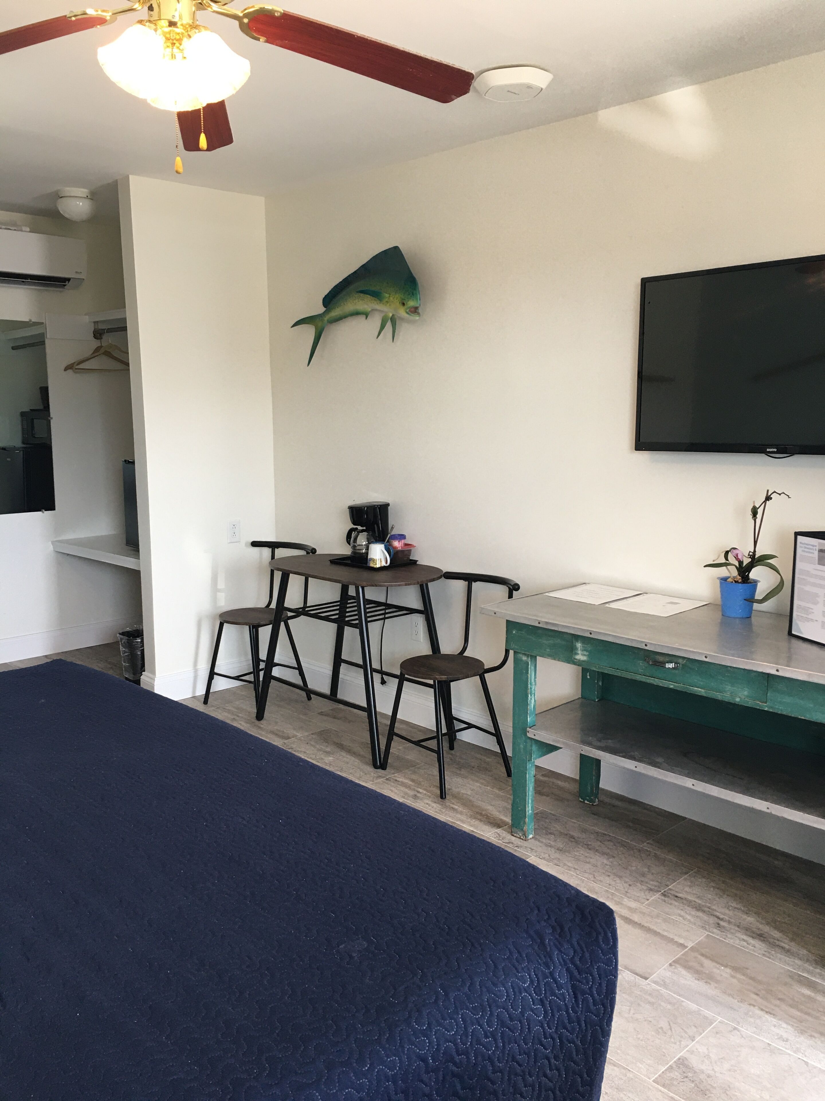 A room with a bed, a ceiling fan, a mounted TV, a desk with a chair, a potted plant, and a fish decoration on the wall.
