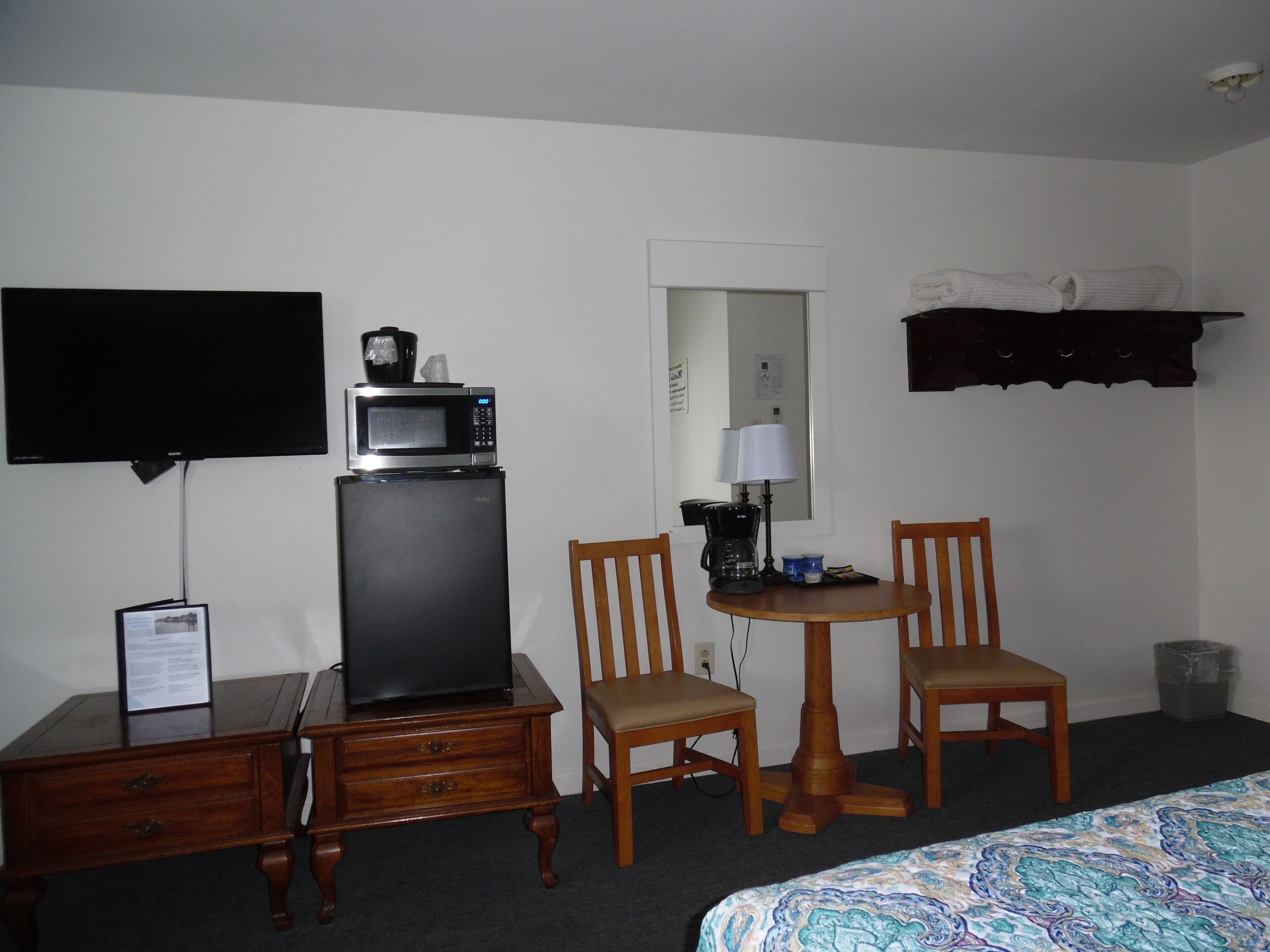A hotel room with a bed, a desk with a television, a microwave, two chairs, a small table with a lamp, and a shelf with a towel.