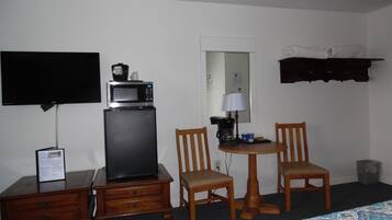 A hotel room with a bed, a desk with a television, a microwave, two chairs, a small table with a lamp, and a shelf with a towel.