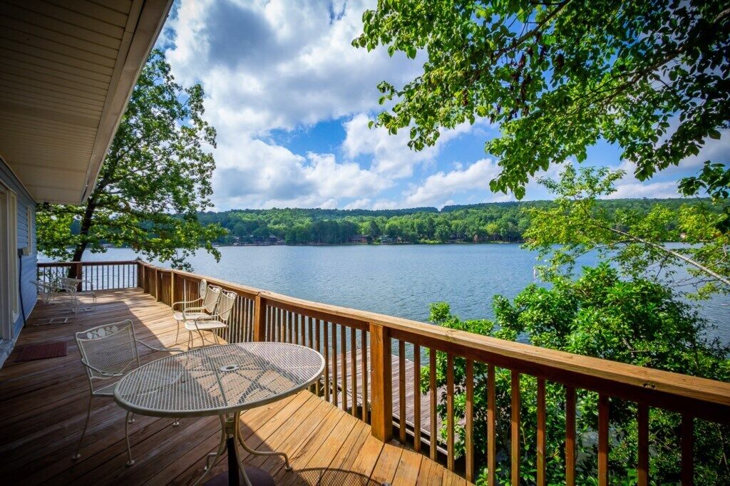 Una terraza de madera con una mesa y sillas, con vistas a un lago rodeado de árboles y colinas.