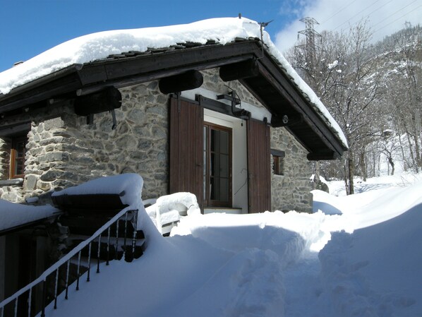 Edificio de piedra con puerta y contraventanas de madera, rodeado de nieve.