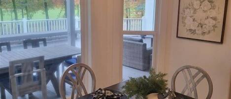 A dining area with a dark wooden table, white chairs, and a chandelier. There is a window with a view of trees and a deck outside.