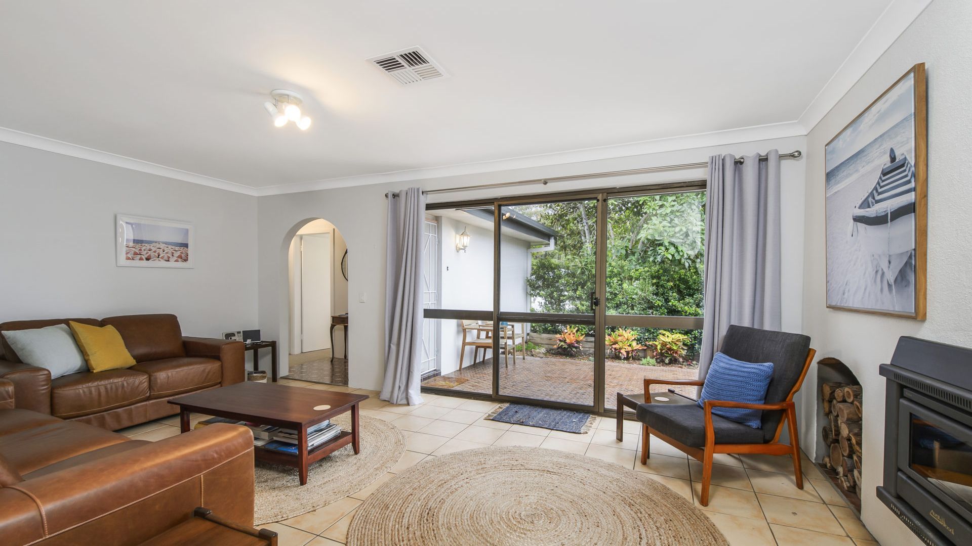 A living room with a sofa, armchair, coffee table, fireplace, and a view through a glass door to an outdoor area with plants.