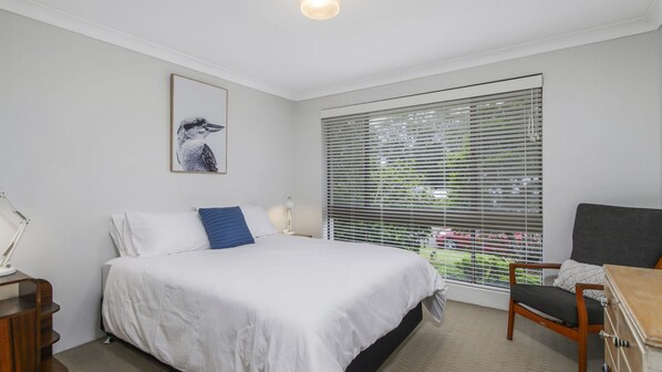 Une chambre d’hôtel avec un lit, une chaise, un bureau, une fenêtre avec des stores et un tableau au mur.