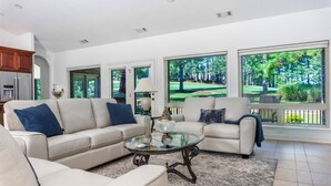 A living room with a beige sectional sofa, a glass coffee table, and large windows offering a view of a green landscape. - 18810 - 10 Cruzado Lane (Hot Springs Village)