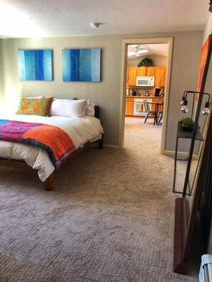 A bedroom with a bed, a carpeted floor, and a view into the kitchen area.