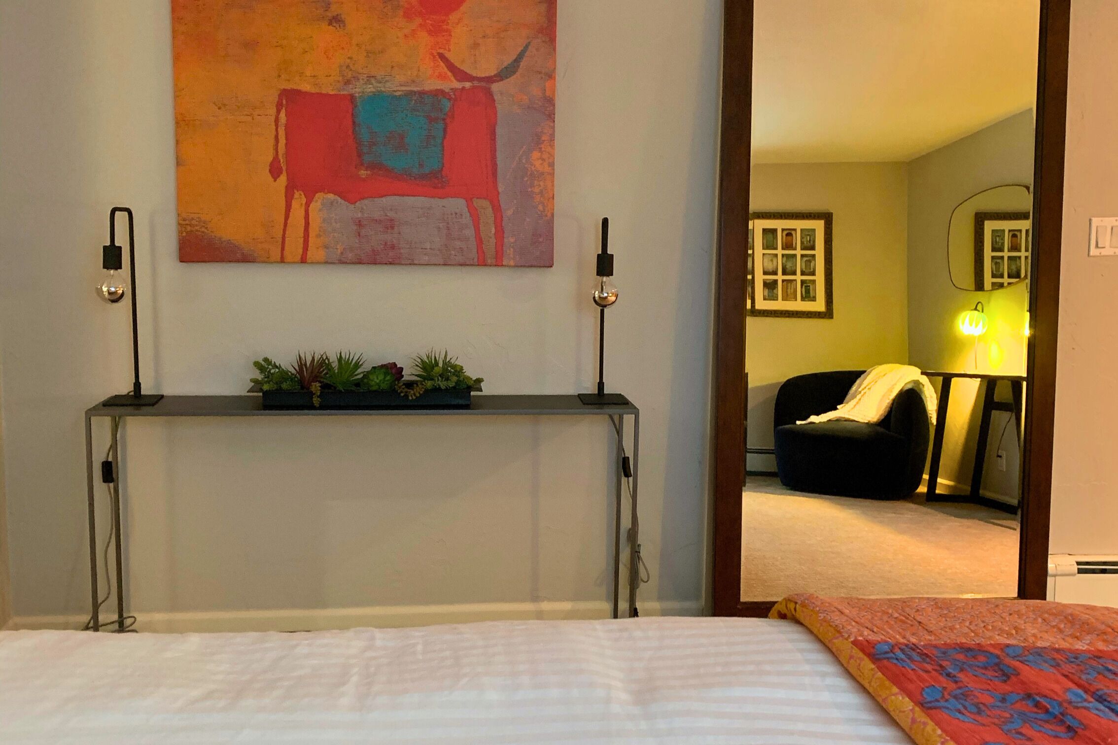 A bedroom with a bed, a bedside table with a plant, a wall-mounted painting, and a door leading to another room.