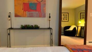 A bedroom with a bed, a bedside table with a plant, a wall-mounted painting, and a door leading to another room.