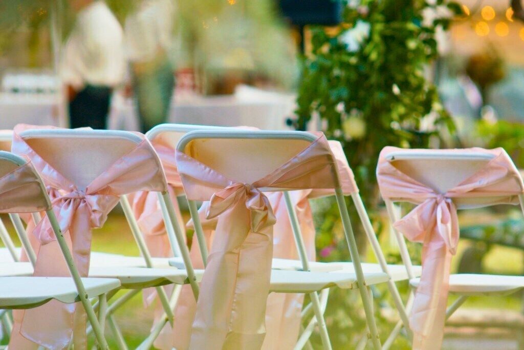 Rows of white chairs with pink ribbons tied at the backrests, arranged outdoors.