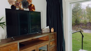 Un salon moderne avec un grand téléviseur à écran plat posé sur un meuble en bois, des œuvres d’art décoratives au mur et une vue sur une rivière et la verdure, visible par une fenêtre.