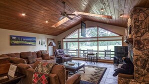 A spacious living room with a high ceiling, large windows, and a stone wall. - 18802 - 2 Granada Way (Hot Springs Village)