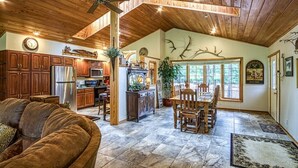 A spacious living area with a wooden ceiling, a dining table with chairs, a kitchen with wooden cabinets, and a large window with a view of trees. - 18802 - 2 Granada Way (Hot Springs Village)