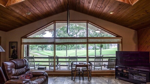 A spacious living room with a large window overlooking a golf course, a leather sofa, a TV, and a fireplace. - 18802 - 2 Granada Way (Hot Springs Village)