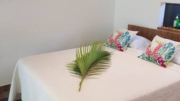 A neatly made bed with a white textured bedspread, a decorative palm leaf, and floral pillows. A wooden stick hangs on the wall above the bed.