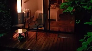 Una habitación acogedora con cama, una mesita y una silla. Cuenta con una puerta corrediza de vidrio que da a un espacio exterior con terraza de madera.