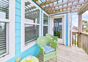 A covered porch with a green wicker chair and a small table, a door leading to a covered area, and a blue exterior wall. - Modern Galveston Beach House! Short Walk to No-Cars Beach. Middle of West End (Galveston)