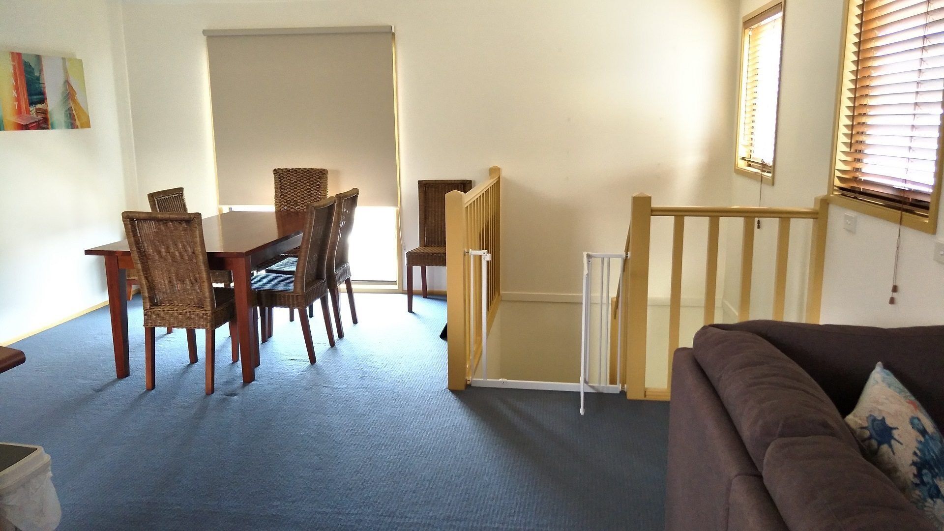 A living room with a brown sofa, a dining table with wicker chairs, a wooden staircase, and a window with blinds.