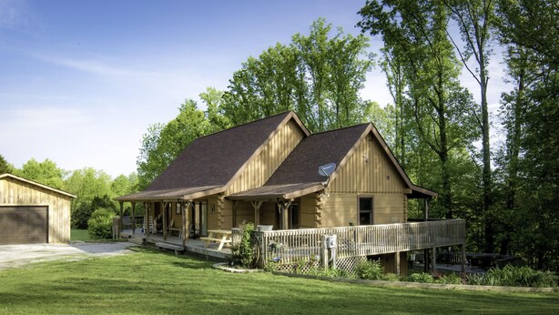 A two-story log cabin with a porch and a deck, surrounded by a well-maintained lawn and trees.