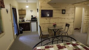 Una habitación acogedora con cama, una pequeña mesa de comedor con sillas, un televisor montado en la pared y una kitchenette con microondas y armarios.