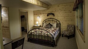 Un dormitorio con estructura de cama metálica, cabecera de madera, una silla, una mesita de noche con lámpara y una puerta que da a otra habitación.