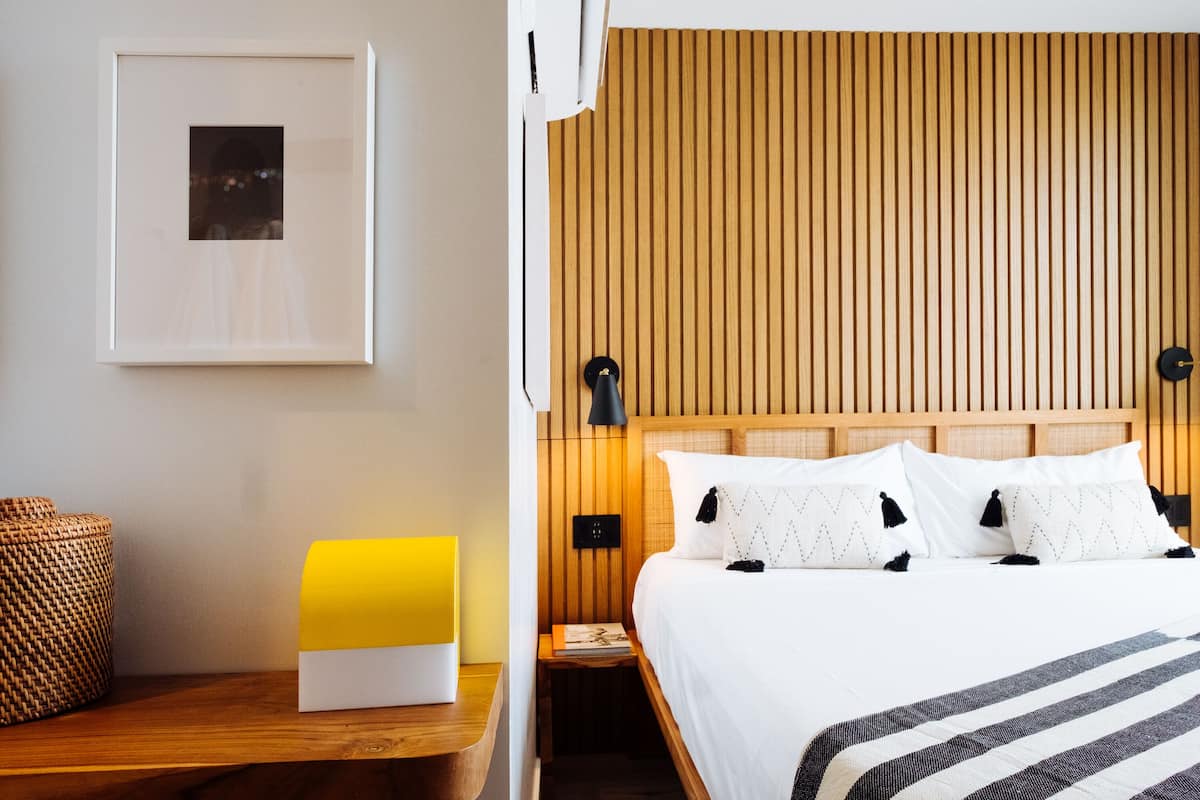 A neatly arranged hotel room with a wooden headboard, a bedside table with a lamp, a framed picture on the wall, and a bed with white bedding and striped throw.
