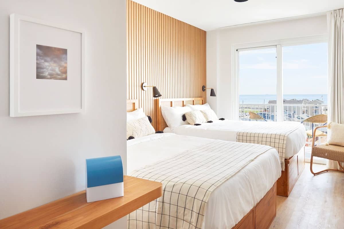 A hotel room with two beds, a large window with a view, a framed picture on the wall, and a small table with a blue object.