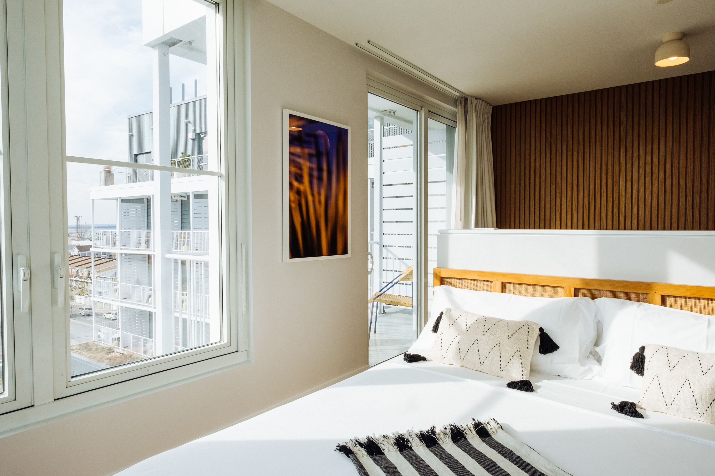 Habitación de hotel con una cama grande, cabecera de madera, vistas de la ciudad a través de amplias ventanas y una obra de arte enmarcada en la pared.