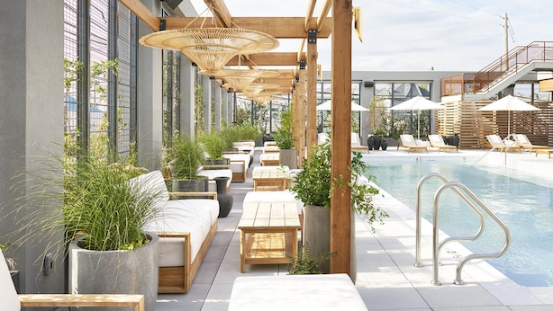 A rooftop pool area with wooden loungers, potted plants, and a view of the cityscape.