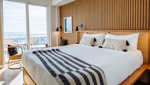 A hotel room with a large bed, wooden headboard, and a view of the city through large windows.