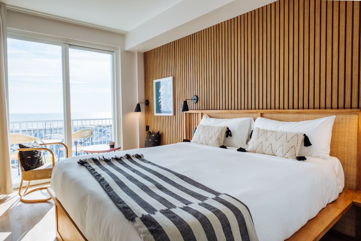 A hotel room with a large bed, wooden headboard, and a view of the city through large windows.
