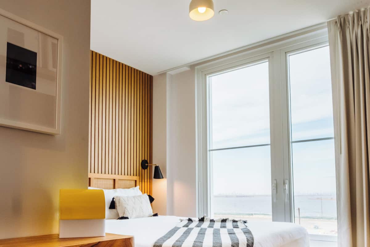 A hotel room with a bed, a bedside table, a lamp, a view of the sea, and a window with curtains.