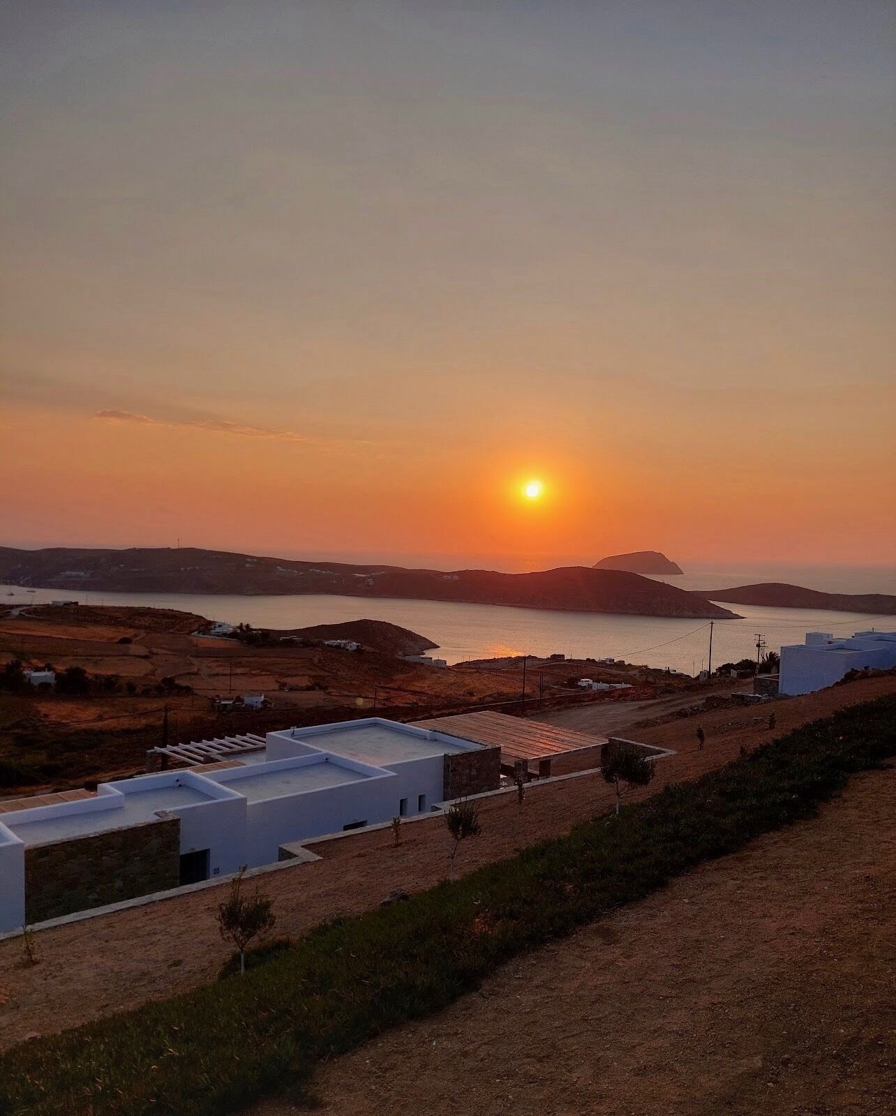 A coastal landscape with a sunset over the sea, featuring a modern building with a flat roof and a small garden area.