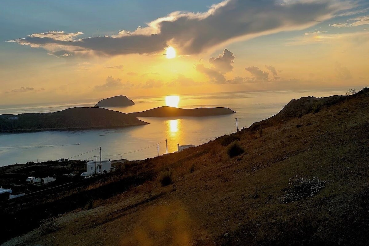 A coastal landscape with a sunset over water, a road leading to a small settlement, and distant hills.