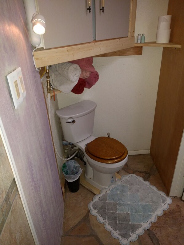 A small bathroom with a toilet, a wooden seat, a towel rack with towels, a shelf with toiletries, and a trash bin. - Little Cottage in Flagstaff (Flagstaff)