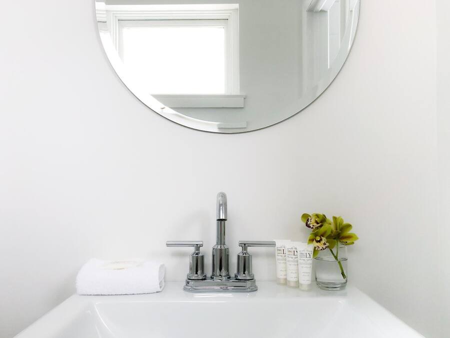A bathroom with a round mirror, a chrome faucet, a white sink, a towel, and a small vase with flowers.